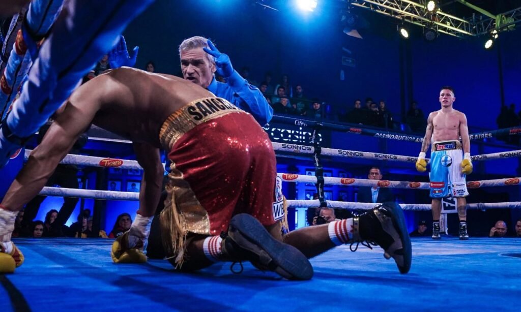 El mexicano Juan Carlos Sánchez López permanece en la lona mientras el árbitro Mario González realiza la cuenta. Al fondo, Alan "El Rusito" Crenz espera la definición tras el golpe que sentenció el combate.